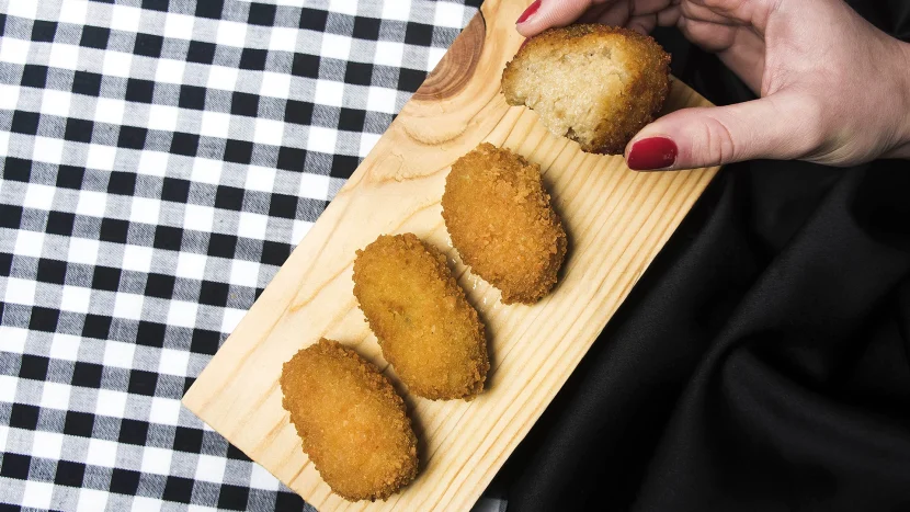 Imagen de varias croquetas de Motril sobre una tabla de madera y una de ellas sostenida con la mano de una mujer